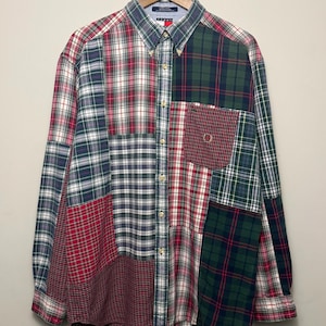 May include: A long-sleeve button-down shirt featuring a patchwork design of various plaid patterns in red, green, white, and navy blue. The shirt includes a chest pocket with a small embroidered logo and a button-down collar on a wooden hanger.
