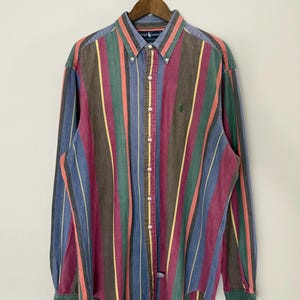 May include: A long-sleeve button-down shirt featuring a vertical striped pattern in shades of green, blue, red, brown, and yellow. The shirt has a button-down collar, a front button placket, and a small embroidered logo on the left chest area.