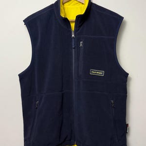 Puede incluir: Un chaleco de forro polar azul marino con forro y cuello de color amarillo brillante. El chaleco cuenta con una cremallera frontal completa, dos bolsillos laterales con cremallera y un bolsillo en el pecho con una etiqueta Polo Sport.