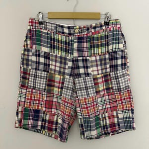 May include: Patchwork shorts featuring a multi-colored plaid pattern with squares of navy blue, red, green, yellow, and white fabric. The shorts have a button closure, a zipper fly, and belt loops, constructed from various tartan fabric swatches.