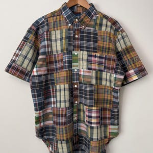 May include: A short-sleeve button-down shirt featuring a multi-colored patchwork design composed of various plaid and tartan patterns in shades of navy, yellow, green, and red. The shirt includes a button-down collar and a small navy blue logo.