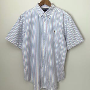 May include: A short-sleeved button-down shirt featuring a vertical striped pattern in light blue, yellow, and white. The shirt has a button-down collar, a front button placket, and a small embroidered Ralph Lauren logo on the left chest area.