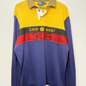 May include: A long-sleeve rugby shirt featuring a color-blocked design with a yellow upper section, a dark green stripe, a red stripe, and a blue lower section. It has a tan collar, a white button placket, and the text 2ND REG with a crown icon.
