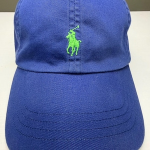 May include: A royal blue baseball cap featuring a bright lime green embroidered logo of a polo player on a horse centered on the front panel. The cap has a curved brim with visible stitching and a structured crown with eyelet ventilation holes.