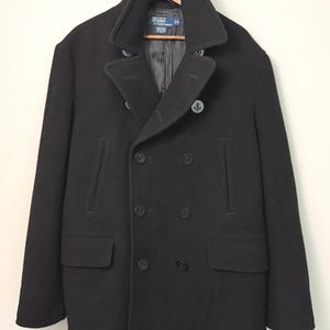 May include: A black double-breasted pea coat featuring a wide notched collar, two vertical slit pockets at the chest, two flap pockets at the waist, and dark buttons embossed with an anchor design. The interior label reads Polo by Ralph Lauren.