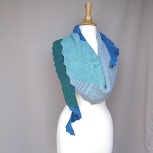 Puede incluir: Una bufanda de punto de color verde azulado, azul y azul claro con un borde festoneado. La bufanda está drapeada sobre un maniquí blanco.