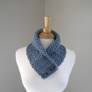 May include: A blue crocheted cowl scarf with two wooden buttons. The scarf is displayed on a white mannequin. The scarf is a solid blue color and appears to be handmade. The cowl scarf is a neck warmer.