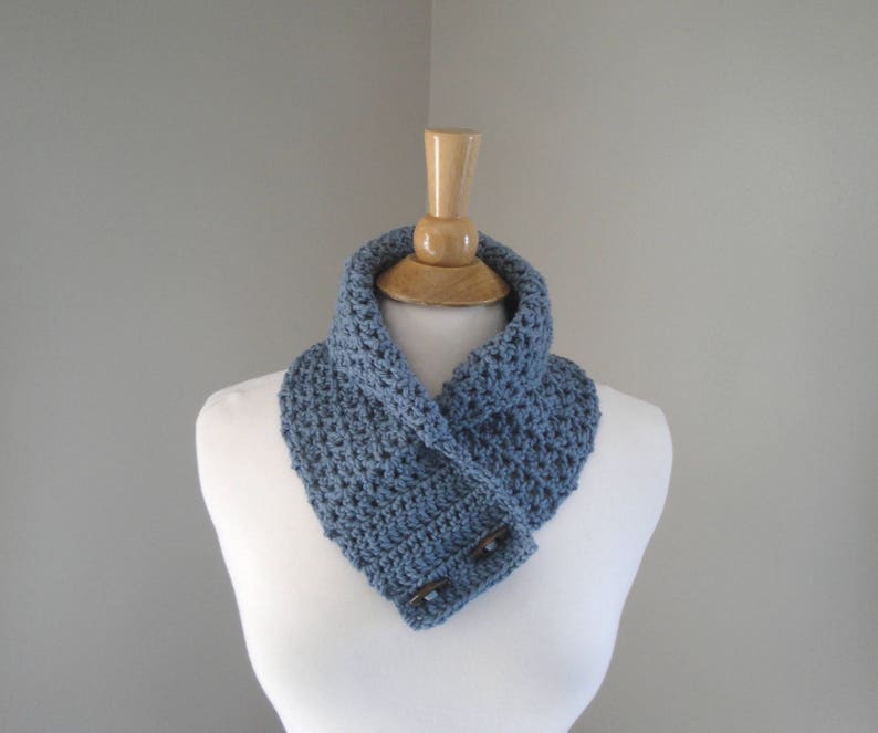 May include: A blue crocheted cowl scarf with two wooden buttons. The scarf is displayed on a white mannequin. The scarf is a solid blue color and appears to be handmade. The cowl scarf is a neck warmer.