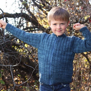 May include: A blue knitted zip-up sweater with a ribbed texture. The sweater has a full zipper and long sleeves. The image shows the sweater on a person in an outdoor setting with a background of trees and branches.