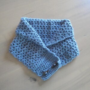 May include: A blue crocheted cowl scarf with a button closure. The scarf is folded over and has a textured appearance. The buttons are a dark brown color. The scarf is made of a soft, knitted material.
