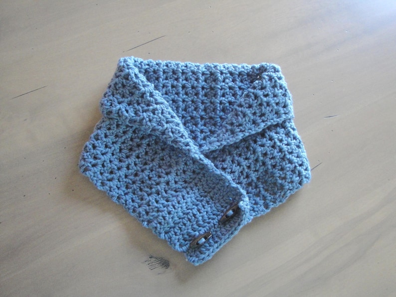May include: A blue crocheted cowl scarf with a button closure. The scarf is folded over and has a textured appearance. The buttons are a dark brown color. The scarf is made of a soft, knitted material.