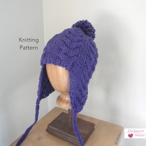 Könnte beinhalten: Lilafarbener Strickhut mit Ohrenklappen und einem Bommel oben. Der Hut wird auf einem Holzständer präsentiert. Strickmuster.