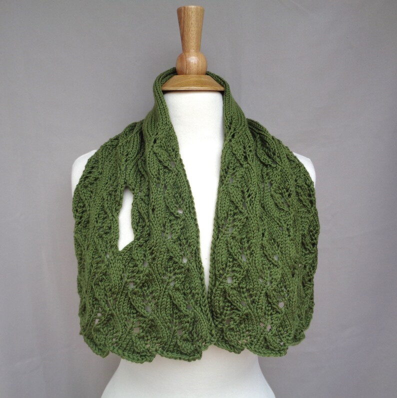 Pode incluir: Uma echarpe de malha verde com um padr&atilde;o de malha de cabo e um delicado design de renda. A echarpe &eacute; exibida em um manequim de madeira.