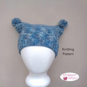 Peut inclure: Un bonnet bleu tricoté avec deux pompons sur le dessus. Le bonnet est sur une tête de mannequin blanche. Le texte "Knitting Pattern" est écrit sous le bonnet. Le texte "Girlpower Designs" est écrit dans un cœur rose sous le texte "Knitting Pattern".