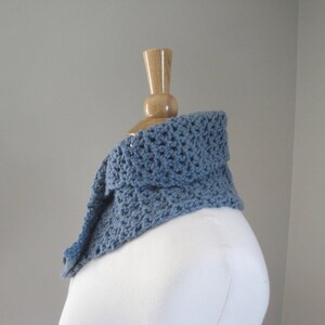 May include: A blue crocheted cowl scarf displayed on a mannequin. The scarf is a soft, muted blue color and has a textured, open weave design. The cowl is folded over, creating a layered effect. The background is a plain, light gray.