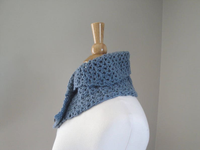 May include: A blue crocheted cowl scarf displayed on a mannequin. The scarf is a soft, muted blue color and has a textured, open weave design. The cowl is folded over, creating a layered effect. The background is a plain, light gray.