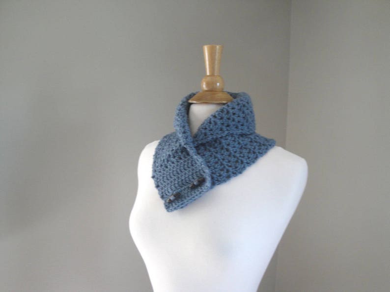 May include: A blue crochet cowl scarf with a button closure, displayed on a white mannequin. The scarf is a solid blue color and appears to be handmade. The cowl is designed to be worn around the neck.
