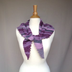 May include: A handmade crochet scarf in shades of purple and gray, displayed on a mannequin. The scarf features a striped pattern and a scalloped edge. The scarf is tied in a triangle shape.