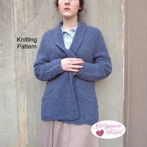 Puede incluir: Un cárdigan azul de punto con cuello chal y bolsillos. El cárdigan está abierto por delante y la modelo lo lleva con una camisa de estampado floral debajo. El texto "Knitting Pattern" es visible en la esquina superior izquierda de la imagen. El texto "Girlpower Designs" es visible en la esquina inferior derecha de la imagen.