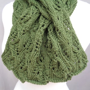 Pode incluir: Uma echarpe de malha verde com um padr&atilde;o de malha de cabo. A echarpe est&aacute; dobrada ao meio e &eacute; exibida em um manequim branco.