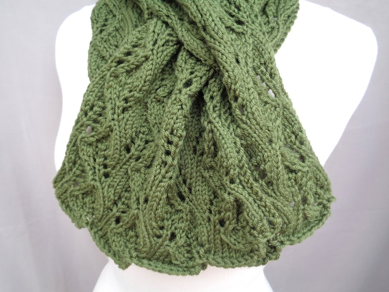 Pode incluir: Uma echarpe de malha verde com um padr&atilde;o de malha de cabo. A echarpe est&aacute; dobrada ao meio e &eacute; exibida em um manequim branco.