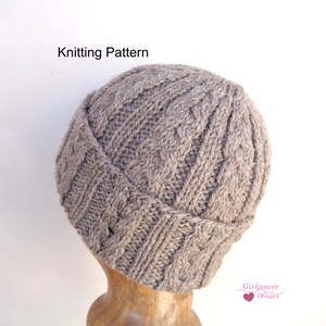 May include: A brown knitted hat with a cable knit pattern. The hat is on a wooden display stand. Knitting Pattern.