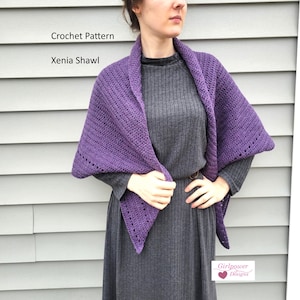 May include: A purple crochet shawl, the "Xenia Shawl", is draped over the shoulders. The shawl is a deep purple color and has a textured stitch pattern. The text "Crochet Pattern" and "Xenia Shawl" are visible in the image.
