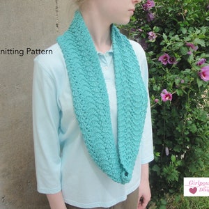 May include: A teal blue knitted cowl neck scarf worn by a person standing in front of a gray wall and a green bush with pink flowers. The text "Knitting Pattern" is visible in the top left corner of the image. The text "Girlpower Designs" is visible in the bottom right corner of the image.