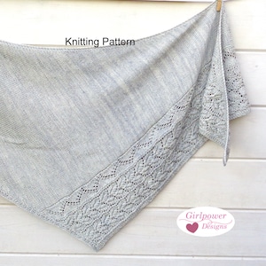 Può includere: Un scialle grigio lavorato a maglia con un bordo in pizzo. Lo scialle è drappeggiato su una superficie di legno bianco. Il testo "Knitting Pattern" è visibile nella parte superiore dell'immagine. Il testo "Girlpower Designs" è visibile nella parte inferiore dell'immagine.