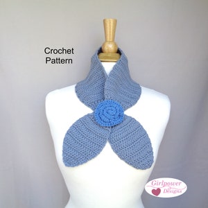 May include: A crocheted scarf in a soft blue hue, featuring a central flower detail in a contrasting blue. The scarf is displayed on a mannequin, showcasing its design and texture. The text "Crochet Pattern" is visible.