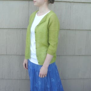 Può includere: Una donna indossa un cardigan verde chiaro con una maglietta bianca sotto e una gonna floreale blu.