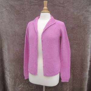 May include: A light pink knitted cardigan with long sleeves and a collar, displayed on a mannequin. The open front features small, matching buttons. The cardigan is made of a soft, textured yarn.