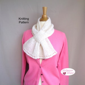 May include: White knitted scarf with a delicate lace trim, tied in a bow around a mannequin wearing a pink cardigan. The text "Knitting Pattern" is visible above the scarf. The text "Girlpower Designs" is visible below the scarf.