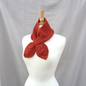 Puede incluir: Bufanda de punto color óxido con un patrón texturizado y detalles de punto trenzado. La bufanda se exhibe en un maniquí blanco, mostrando su diseño y forma. Un accesorio cálido.