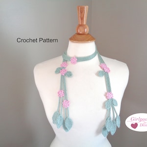 Könnte beinhalten: Ein gehäkelter Schal in einem sanften Grünton, verziert mit rosa Blüten und grünen Blättern. Der Schal wird auf einer Schaufensterpuppe präsentiert, die das zarte Blumendesign hervorhebt. Der Text "Crochet Pattern" ist sichtbar.