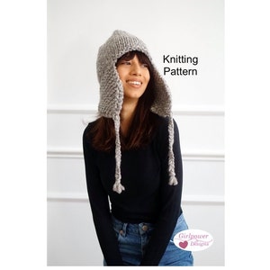 Puede incluir: Un gorro de punto gris con lazos largos. El gorro tiene una capucha puntiaguda y se combina con una camisa negra y unos vaqueros azules. El texto "Knitting Pattern" es visible en la imagen.