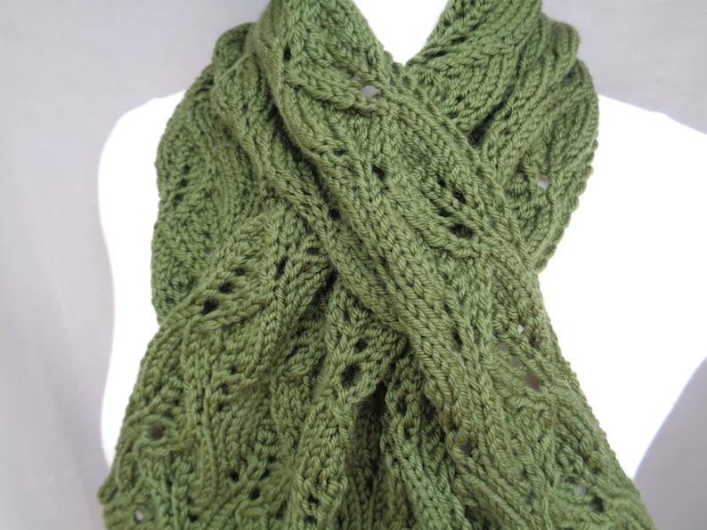 Pode incluir: Uma echarpe de malha verde com um padr&atilde;o de malha de cabo. A echarpe est&aacute; dobrada ao meio e drapeada sobre o pesco&ccedil;o de um manequim.