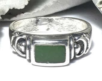 Puede incluir: Un anillo de plata con una piedra verde engastada en un marco cuadrado. El anillo tiene un diseño decorativo en la banda.