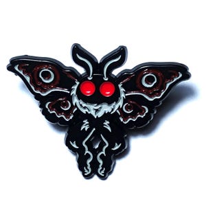 Puede incluir: Pin de esmalte negro de una polilla con ojos rojos y alas marrones y negras. La polilla está representada en un estilo de dibujos animados.