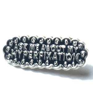 May include: A silver-colored pin with a black background. The pin has a cloud-like border and says "Ask me about my hyperfixations".