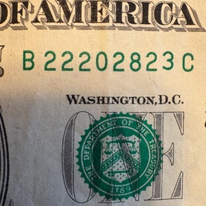 May include: Close-up of a United States one-dollar bill. The bill features the text "OF AMERICA" and a green seal with the words "DEPARTMENT OF THE TREASURY". The serial number "B 22202823 C" is printed in green.