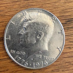 May include: Silver US half dollar coin featuring a profile of John F. Kennedy with the inscription "In God We Trust" and the dates "1776-1976".