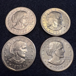 May include: Four Susan B. Anthony dollar coins. Each coin features a profile of Susan B. Anthony, the word "LIBERTY", and the phrase "IN GOD WE TRUST". Two coins are dated 1979, and two are dated 1980.