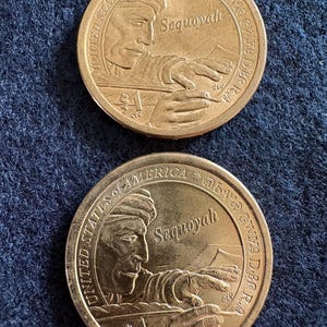 May include: Two gold-colored United States of America dollar coins. Each coin features a profile of Sequoyah, the Cherokee silversmith, and the inscription "Sequoyah". The coins are on a dark blue textured surface.