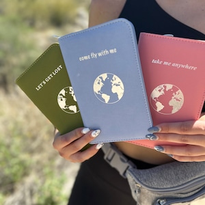 May include: Three travel passport holders in olive green, light blue, and pink. Each passport holder features a world map graphic and a travel-themed phrase: "Let's Get Lost," "Come Fly With Me," and "Take Me Anywhere."