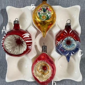 May include: Four vintage Christmas ornaments in a white ceramic holder. The ornaments are red, gold, and blue with silver and white accents. Each ornament has a metal cap for hanging. The ornaments have unique shapes and designs.
