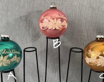 May include: Three vintage Christmas ornaments in teal, pink, and gold, each with the words "Merry Christmas" and a floral design. The ornaments are displayed on black metal stands, perfect for holiday decor.