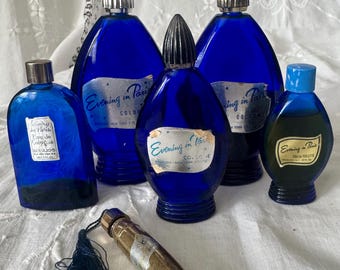 May include: A collection of five cobalt blue glass perfume bottles labeled Evening in Paris. Four bottles feature silver-toned caps, while one smaller bottle has a light blue cap. A slender, metallic, cylindrical perfume vial with a black tassel is included.