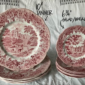 May include: A set of pink and white ceramic plates featuring a castle and bridge scene with floral borders. The larger plates are labeled 10 inch dinner plates, and the smaller plates are labeled 6 3/4 inch salad or bread plates.