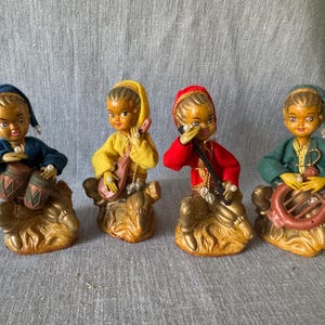 May include: Four vintage figurines of musicians. Each figure is dressed in a different color outfit: blue, yellow, red, and green. They are playing drums, a lute, a wind instrument, and a harp. The figurines are on a gold-colored base.